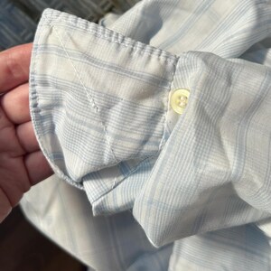 May include: A long-sleeved shirt cuff featuring a light blue and white striped pattern. The cuff is constructed with a single white button and visible stitching along the edges, showcasing the woven fabric texture and the classic design of the sleeve.