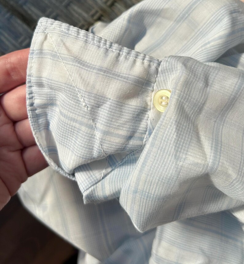May include: A long-sleeved shirt cuff featuring a light blue and white striped pattern. The cuff is constructed with a single white button and visible stitching along the edges, showcasing the woven fabric texture and the classic design of the sleeve.