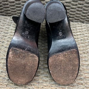 May include: A pair of black leather ankle boots with block heels. The soles are worn, showing signs of use. The heels are approximately 3 inches high. The boots are set against a woven background.