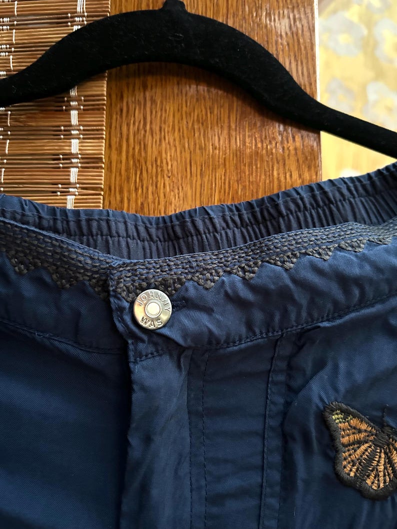May include: Close-up of navy blue shorts with an embroidered waistband and button closure. The shorts have a small butterfly patch. The silver button is printed with the words "Johnny Was".