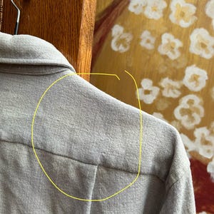 以下が含まれることがあります： 明るい灰色のシャツのクローズアップ。背中のヨークと襟を示しています。シャツは金属製のハンガーに掛けられています。背景には木製の表面と、白と金のフローラルデザインの絵画が含まれています。