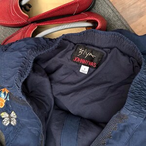 May include: Navy blue Johnny Was top with embroidered butterfly and floral details. The top features a gathered neckline and the brand's label. Red leather shoes are visible in the background.