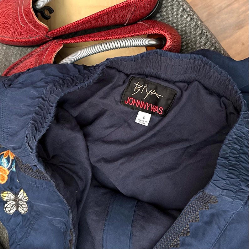May include: Navy blue Johnny Was top with embroidered butterfly and floral details. The top features a gathered neckline and the brand's label. Red leather shoes are visible in the background.