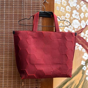 May include: A burgundy tote bag with a geometric honeycomb pattern. The bag has matching burgundy handles and trim. The bag is hanging on a black hanger against a bamboo blind and a painted floral background.