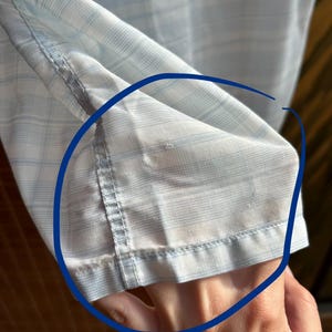 May include: A light blue and white plaid shirt sleeve featuring a visible area of fabric wear and fraying along the hemline and stitching, highlighted by a blue circular marking on the garment fabric.