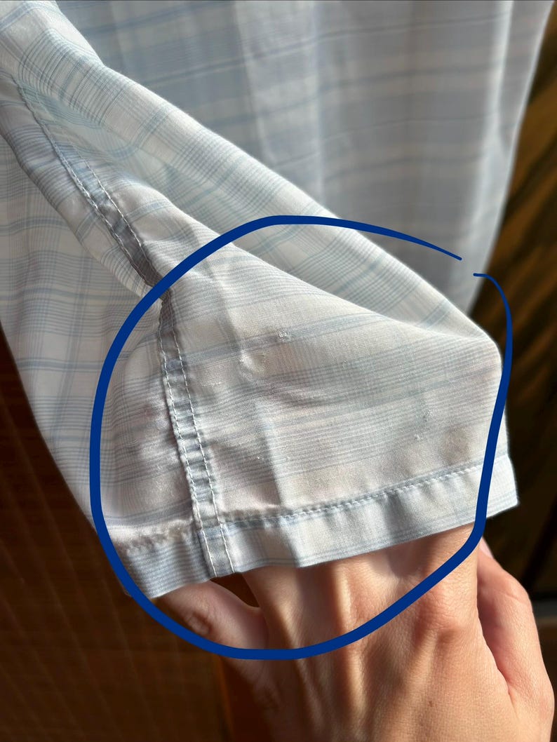 May include: A light blue and white plaid shirt sleeve featuring a visible area of fabric wear and fraying along the hemline and stitching, highlighted by a blue circular marking on the garment fabric.