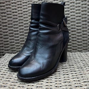 May include: A pair of black leather ankle boots with a block heel. The boots have a rounded toe, a buckle detail on the side, and a textured appearance. The boots are displayed on a woven surface.