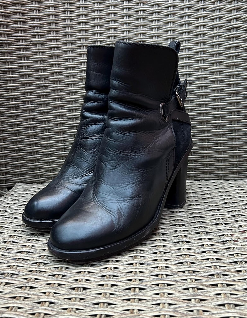 May include: A pair of black leather ankle boots with a block heel. The boots have a rounded toe, a buckle detail on the side, and a textured appearance. The boots are displayed on a woven surface.