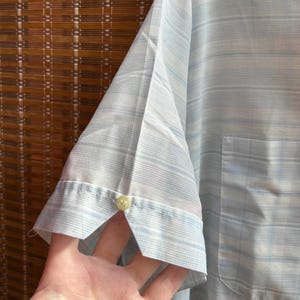 May include: A short-sleeved button-down shirt in a light blue and white striped pattern. The sleeve features a small notched cuff detail secured with a single light-colored button. The fabric has a fine, semi-sheer texture with a chest pocket.