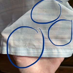 May include: A light blue and white striped fabric garment featuring three blue circles drawn on the surface to highlight small areas of discoloration and a tiny hole near the bottom hem of the material.