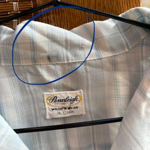 May include: A light blue and white plaid button-down shirt with a collar. A white fabric label is stitched at the neck, featuring the text Pennleigh, WASH 'N WEAR, and 16 L 16 1/2. A small dark stain is visible on the inner collar fabric.
