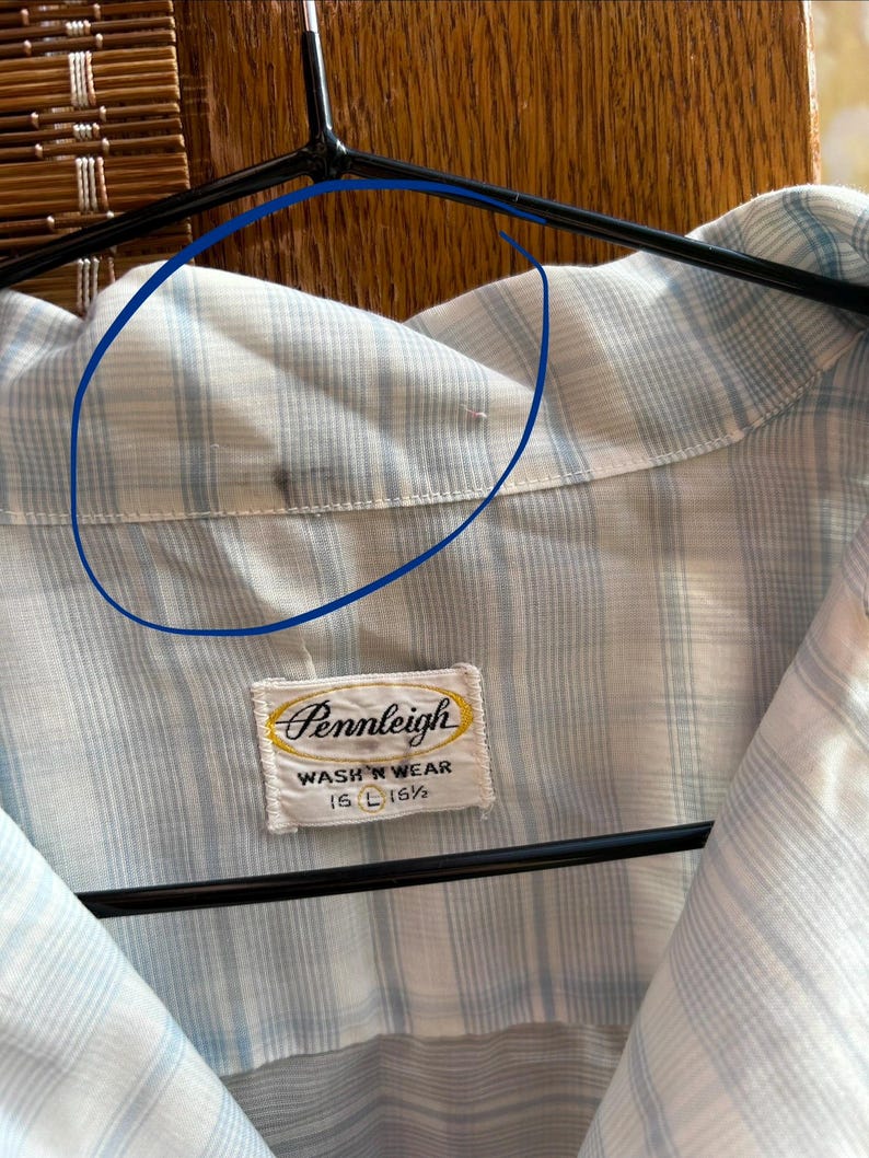 May include: A light blue and white plaid button-down shirt with a collar. A white fabric label is stitched at the neck, featuring the text Pennleigh, WASH 'N WEAR, and 16 L 16 1/2. A small dark stain is visible on the inner collar fabric.