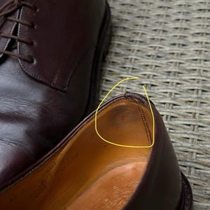 May include: Close-up of a pair of dark brown leather dress shoes. One shoe is in focus, revealing the interior and stitching details. The other shoe is partially visible, with laces tied. The shoes are on a textured surface.