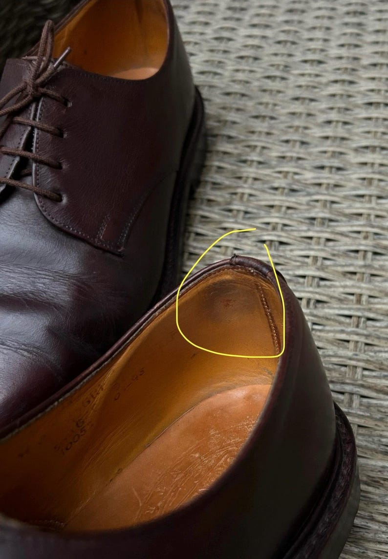 May include: Close-up of a pair of dark brown leather dress shoes. One shoe is in focus, revealing the interior and stitching details. The other shoe is partially visible, with laces tied. The shoes are on a textured surface.