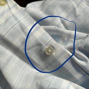 May include: A light blue and white plaid button-down shirt featuring a circular blue ink mark drawn around a single translucent plastic button on the front placket. The fabric displays a fine grid pattern of intersecting light blue lines.