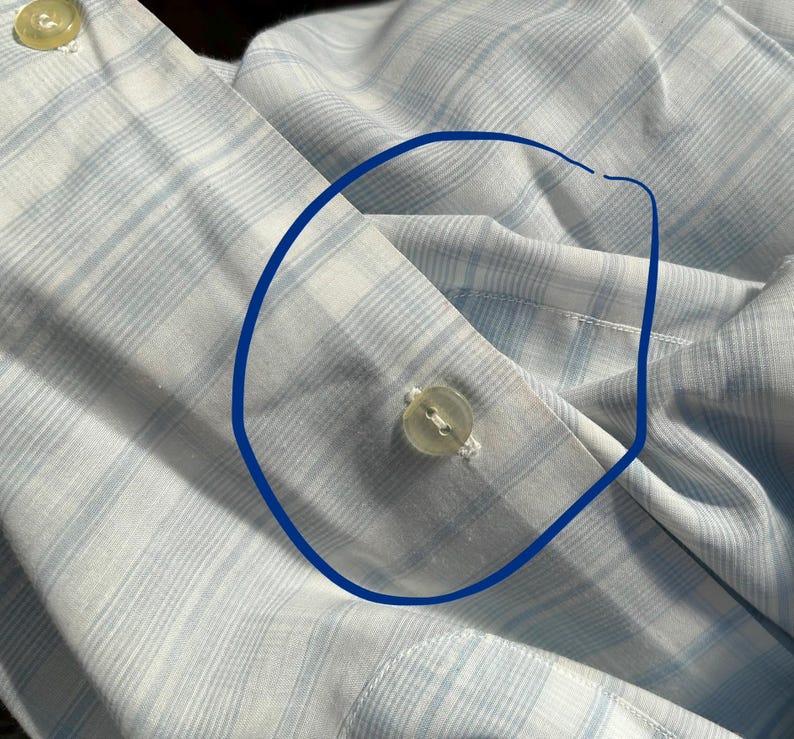 May include: A light blue and white plaid button-down shirt featuring a circular blue ink mark drawn around a single translucent plastic button on the front placket. The fabric displays a fine grid pattern of intersecting light blue lines.