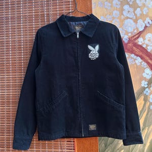 May include: Black corduroy jacket with a collared neckline, a zipper closure, and two front pockets. A patch featuring a rabbit skull is on the chest. The jacket has the text "SSDD" on a label near the bottom and on the collar.