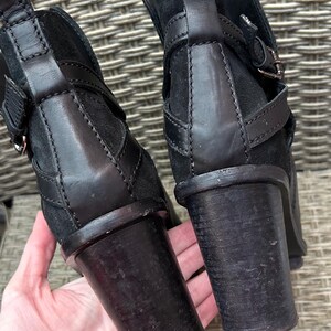 May include: Close-up of two black ankle boots with stacked heels. The boots feature leather straps with silver buckles and a suede upper. The heels are approximately 3 inches tall.