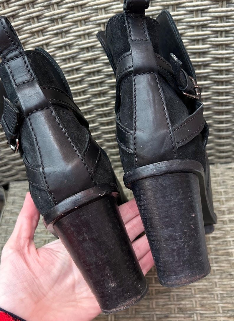 May include: Close-up of two black ankle boots with stacked heels. The boots feature leather straps with silver buckles and a suede upper. The heels are approximately 3 inches tall.