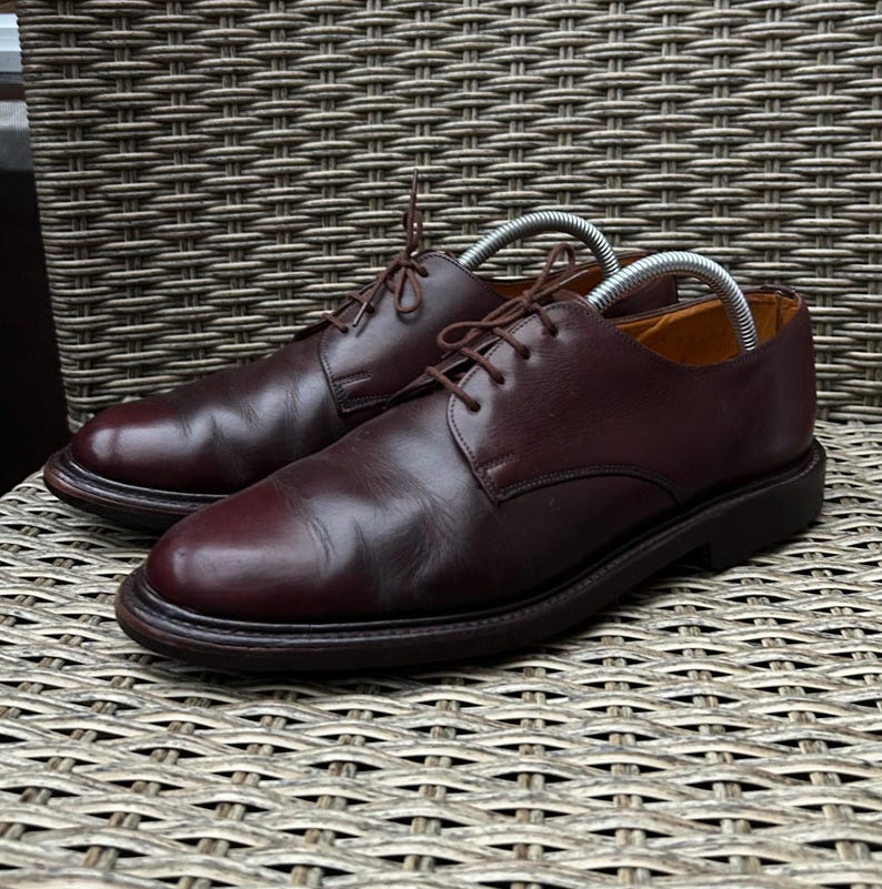 May include: A pair of burgundy leather dress shoes with laces. The shoes have a classic design with a rounded toe and a low heel. Shoe trees are inserted in the shoes. The shoes are placed on a woven surface.