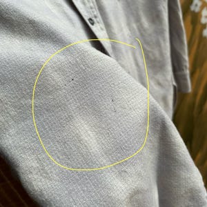 以下が含まれることがあります： 明るいグレーのボタンダウンシャツのクローズアップ。シャツには胸ポケットが2つとクラシックな襟が付いています。生地は、織り込まれたテクスチャード加工の素材のようです。シャツはボタンが外されており、衣服の内側が見えています。