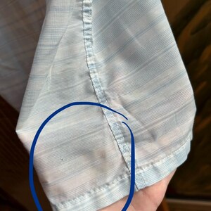 May include: A light blue and white striped button-down shirt sleeve featuring a vertical seam. A small, dark circular mark is visible on the fabric within the blue-circled area, indicating a minor imperfection on the light-colored material.