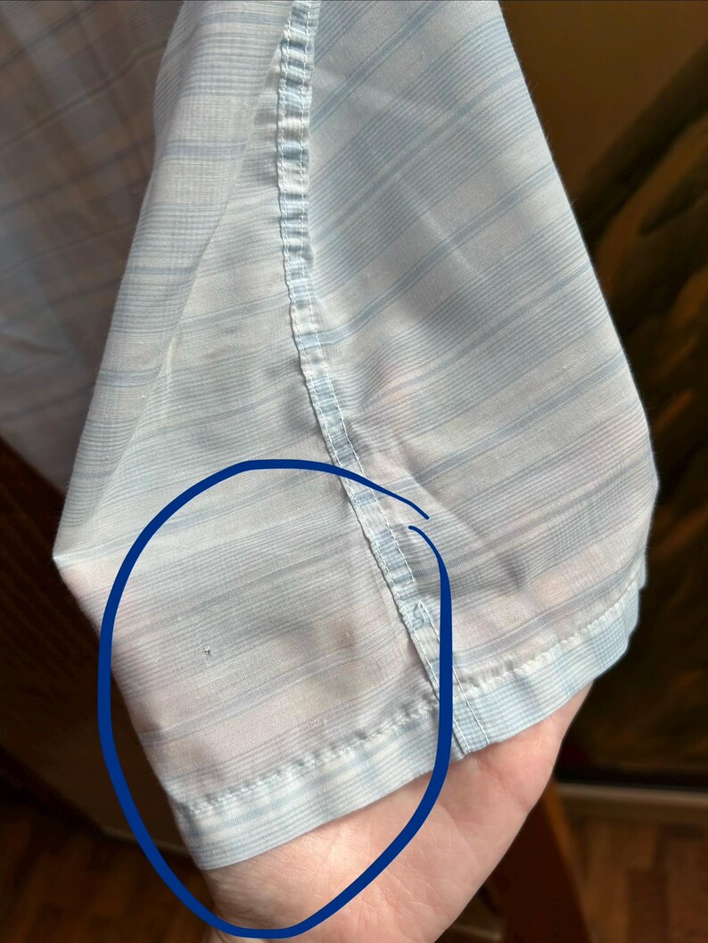 May include: A light blue and white striped button-down shirt sleeve featuring a vertical seam. A small, dark circular mark is visible on the fabric within the blue-circled area, indicating a minor imperfection on the light-colored material.