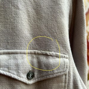 以下が含まれることがあります： ベージュのボタンダウンシャツのクローズアップ。 シャツには、ボタン留めのシングルポケットが付いています。 生地は質感のある外観です。