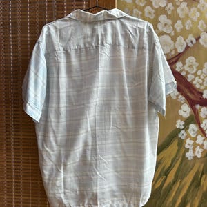 May include: A short-sleeved button-down shirt featuring a light blue and white horizontal striped pattern. The shirt has a classic collar, a straight hemline, and is displayed hanging on a metal hanger against a textured background.