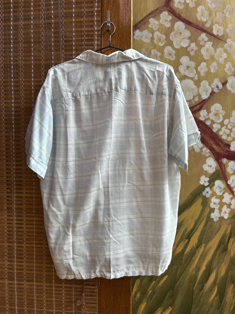 May include: A short-sleeved button-down shirt featuring a light blue and white horizontal striped pattern. The shirt has a classic collar, a straight hemline, and is displayed hanging on a metal hanger against a textured background.