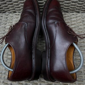 May include: A pair of dark brown leather dress shoes with laces. The shoes are placed side by side, with shoe trees inserted. The shoes are on a woven surface.
