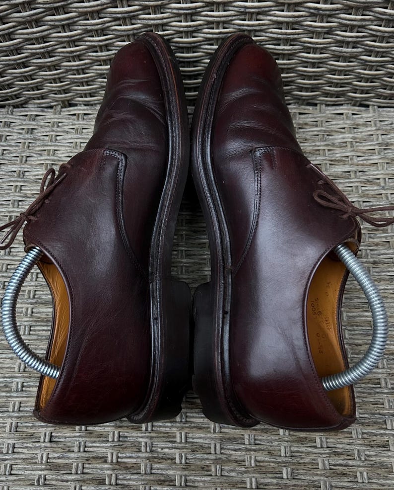 May include: A pair of dark brown leather dress shoes with laces. The shoes are placed side by side, with shoe trees inserted. The shoes are on a woven surface.