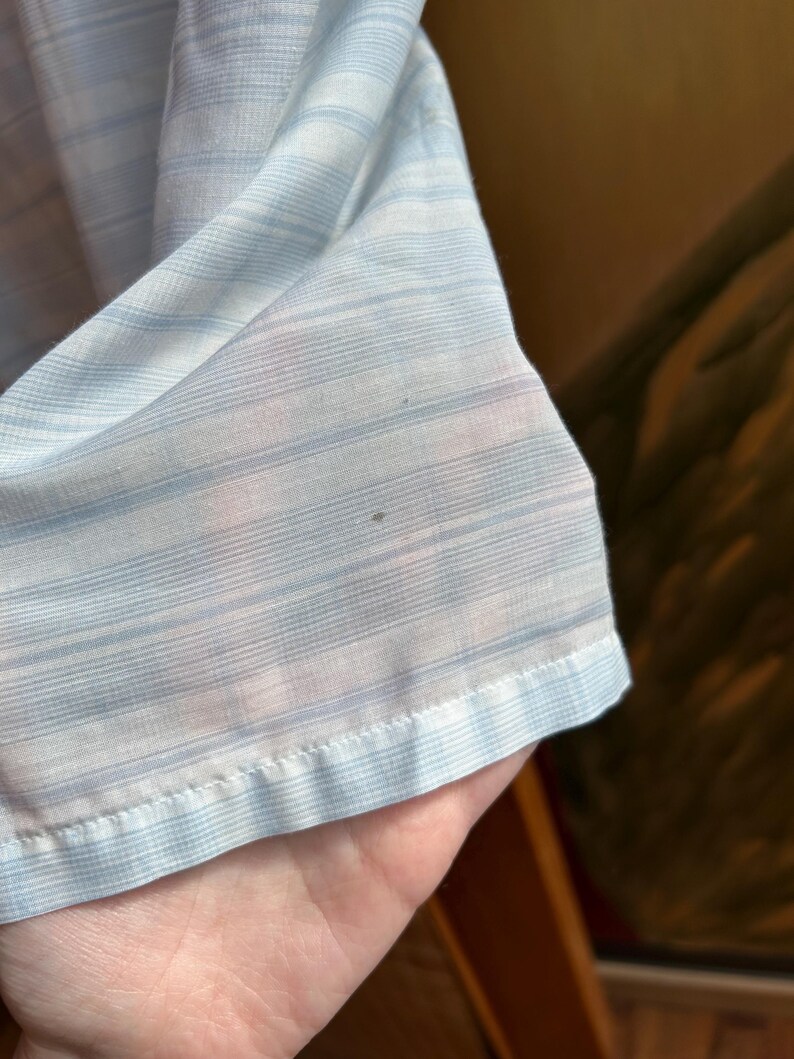 May include: A lightweight, semi-sheer fabric featuring a pattern of thin, horizontal light blue and white stripes. The material has a visible, fine-textured weave and a finished hem along the bottom edge, showing a small dark spot on the surface.