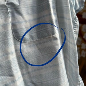 May include: A light blue and white plaid button-down shirt featuring a chest pocket. The fabric displays a subtle grid pattern with horizontal and vertical lines. A blue circular outline is drawn over the pocket area to highlight the specific section.