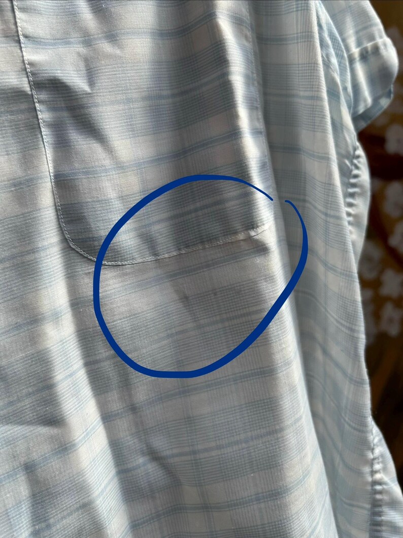 May include: A light blue and white plaid button-down shirt featuring a chest pocket. The fabric displays a subtle grid pattern with horizontal and vertical lines. A blue circular outline is drawn over the pocket area to highlight the specific section.