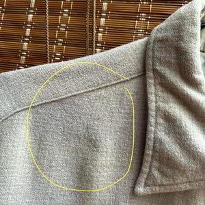 以下が含まれることがあります： 明るいタン色のボタンダウンシャツのクローズアップ。生地は質感のある外観です。襟と肩の縫い目が見え、衣服の仕立てられた構造を示しています。シャツは織り素材で作られているようです。