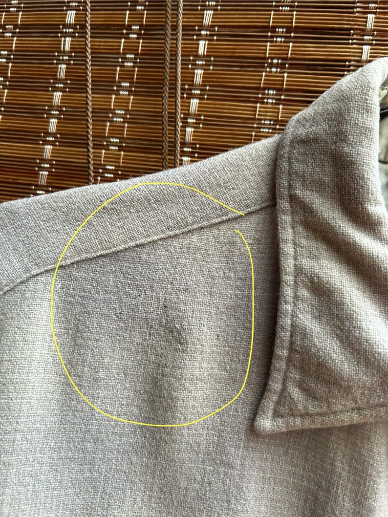 以下が含まれることがあります： 明るいタン色のボタンダウンシャツのクローズアップ。生地は質感のある外観です。襟と肩の縫い目が見え、衣服の仕立てられた構造を示しています。シャツは織り素材で作られているようです。