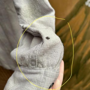 以下が含まれることがあります： 明るい灰色の長袖シャツの袖のクローズアップ。袖口はボタンで留められており、小さな黒い点が見えます。生地は質感のある外観をしています。