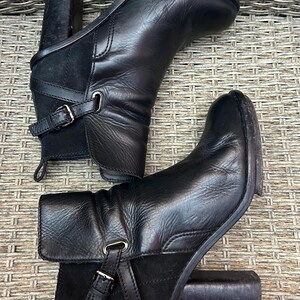 May include: A pair of black leather ankle boots with a block heel. The boots have a buckle detail on the side and a suede panel. The boots are approximately 4 inches high.