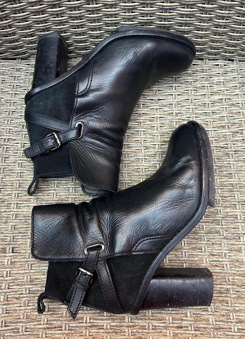 May include: A pair of black leather ankle boots with a block heel. The boots have a buckle detail on the side and a suede panel. The boots are approximately 4 inches high.