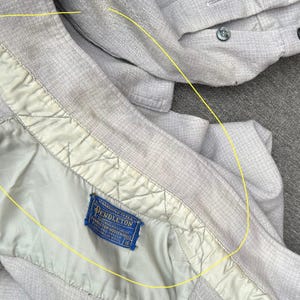 以下が含まれることがあります： 織り目加工が施された、明るいグレーのジャケットのクローズアップ。内側の裏地は明るい緑色で、青色のペンドルトンラベルが見えます。ジャケットのデザインには、クリーム色のトリムが含まれています。