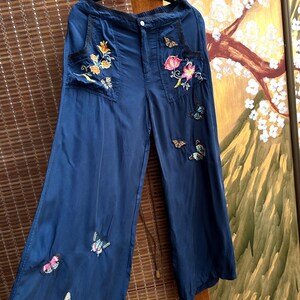 May include: Navy blue wide-leg trousers with embroidered floral and butterfly details. The trousers feature two front pockets with floral embroidery in shades of yellow, orange, and pink. Butterflies in various colours are scattered across the legs.