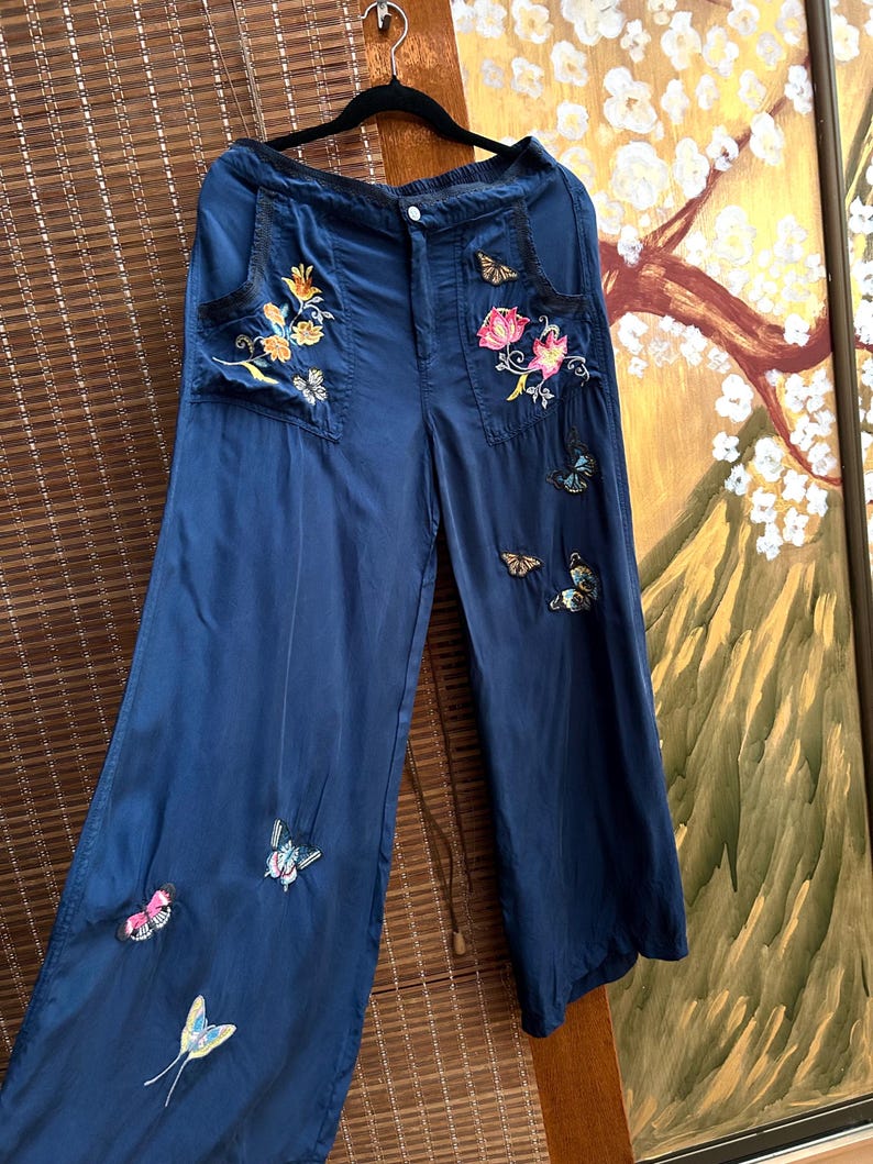 May include: Navy blue wide-leg trousers with embroidered floral and butterfly details. The trousers feature two front pockets with floral embroidery in shades of yellow, orange, and pink. Butterflies in various colours are scattered across the legs.
