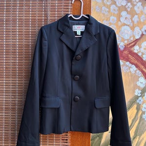 May include: A black blazer with a classic design, featuring a notched lapel, three-button closure, and two front pockets. The blazer is hanging on a pink hanger against a neutral background.