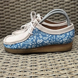 May include: A pair of blue and white paisley patterned shoes with beige suede accents and laces. The shoes have a brown sole and a metal shoe tree inserted inside. The shoes are on a woven surface.