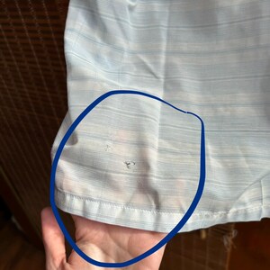 May include: A light blue and white striped shirt featuring a horizontal pattern. The fabric shows two small, distinct holes in the lower section, which are highlighted by a dark blue circular outline drawn directly onto the garment's surface.