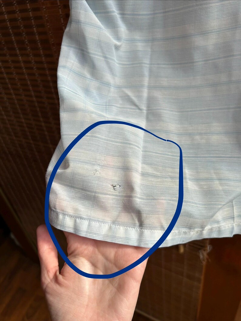 May include: A light blue and white striped shirt featuring a horizontal pattern. The fabric shows two small, distinct holes in the lower section, which are highlighted by a dark blue circular outline drawn directly onto the garment's surface.