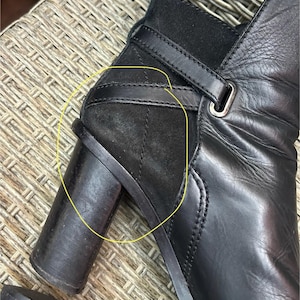 May include: Close-up of a black leather ankle boot with a block heel. The boot features a suede panel on the side, a strap with a buckle, and a rounded toe. The leather appears to be slightly textured.