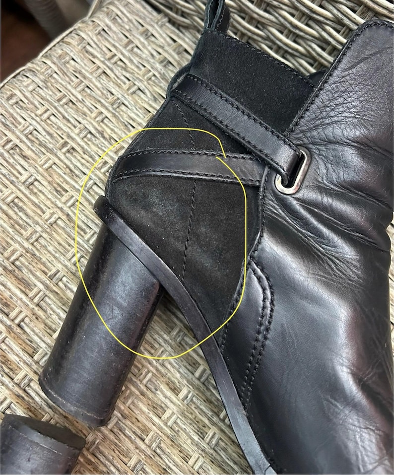 May include: Close-up of a black leather ankle boot with a block heel. The boot features a suede panel on the side, a strap with a buckle, and a rounded toe. The leather appears to be slightly textured.