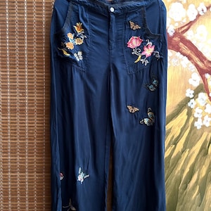 Peut inclure: Pantalon large bleu marine avec broderies florales et papillons. Le pantalon présente des poches ornées de broderies florales colorées et d'appliques papillon. Le tissu semble léger, adapté aux températures plus chaudes.